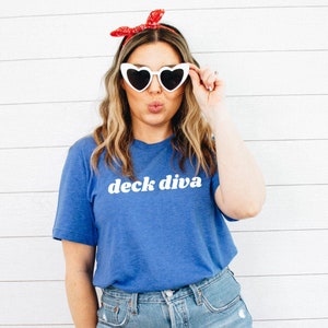 May include: A woman wearing a blue t-shirt with the text "deck diva" in white letters. She is wearing denim shorts, white heart-shaped sunglasses, and a red headband.