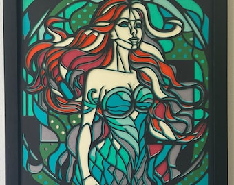 Op de afbeelding: Een kunstwerk van papier-cut-out dat een vrouw met lang rood haar afbeeldt die een blauwe en groene jurk in zeemeermin-stijl draagt. Het kunstwerk is in zwart ingelijst.