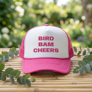 May include: A pink and white trucker hat with the words "BIRD BAM CHEERS" in pink block letters. The hat has a mesh back and a curved brim. The hat is sitting on a wooden surface.