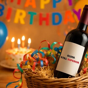 Peut inclure: Une bouteille de vin rouge avec une étiquette blanche portant l'inscription "OLDER. STILL SUPERIOR." est dans un panier en osier avec des serpentins colorés. Un gâteau d'anniversaire avec des bougies allumées et des ballons sont en arrière-plan.
