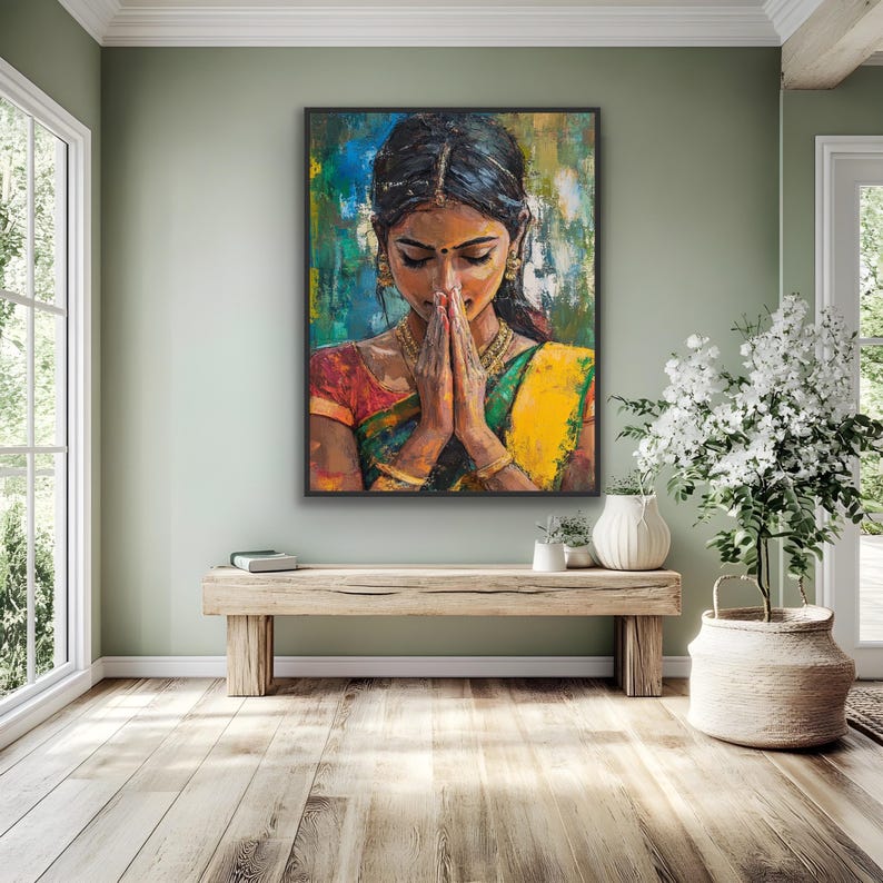 May include: A framed painting depicts a Indian wall art Indian painting woman in traditional Indian attire with hands clasped in prayer. The artwork features a vibrant color palette of yellow, green, red, and blue. The woman is adorned with gold jewelry, and the background has abstract brushstrokes.