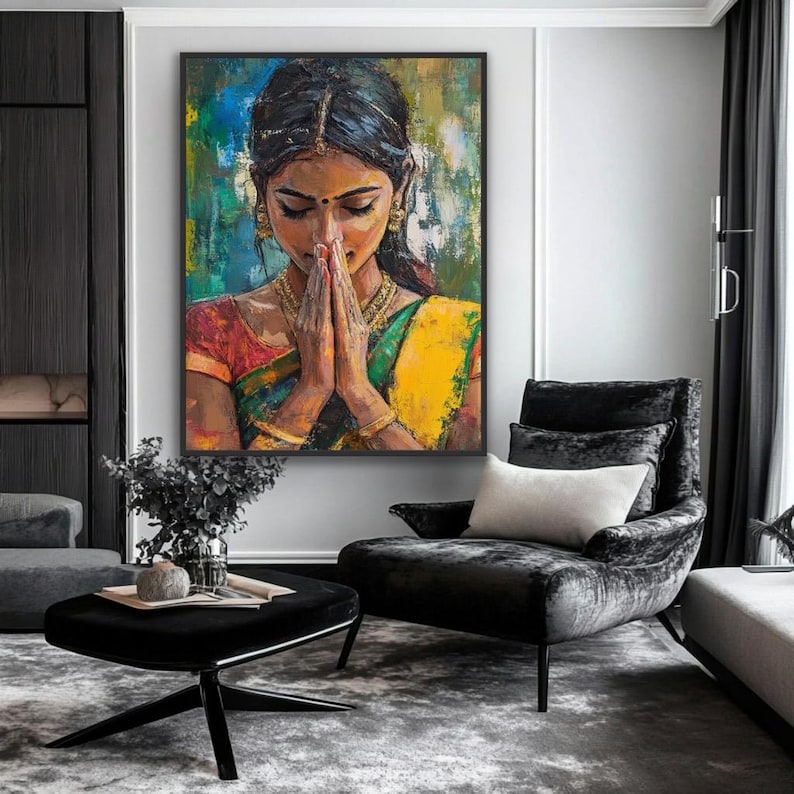 May include: A framed painting depicting a woman in traditional Indian dress, hands in prayer. The artwork uses vibrant colours, including yellow, green, and red, with gold jewellery. The painting hangs above a black armchair in a modern living room setting.