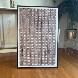 Pode incluir: Uma obra de arte abstrata emoldurada com um padr&atilde;o tecido marrom e branco. A obra retangular tem uma moldura marrom escura e uma borda branca. &Eacute; exibida em um piso de parquet, adicionando um toque decorativo.