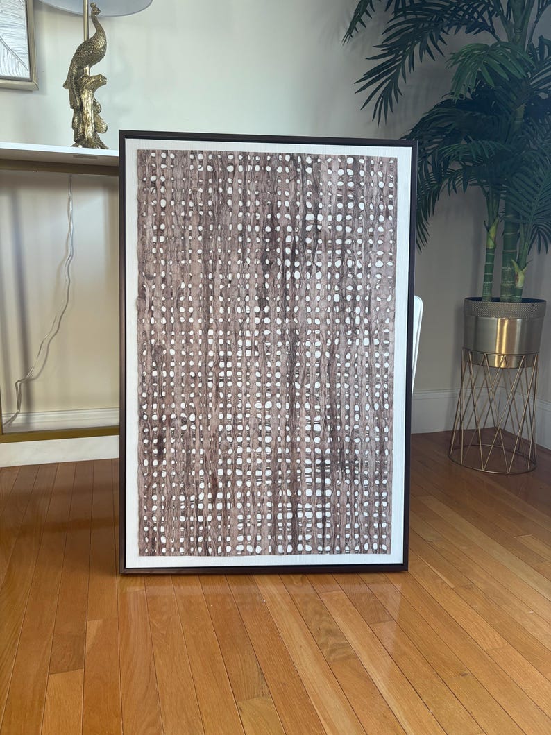 Pode incluir: Uma obra de arte abstrata emoldurada com um padr&atilde;o tecido marrom e branco. A obra retangular tem uma moldura marrom escura e uma borda branca. &Eacute; exibida em um piso de parquet, adicionando um toque decorativo.