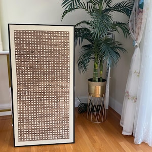 Pode incluir: Uma obra de arte abstrata emoldurada com um padr&atilde;o de pontos marrons e brancos. A obra de arte est&aacute; em uma moldura preta e encostada em uma parede. Uma palmeira alta em vaso e um suporte de planta dourado est&atilde;o ao fundo.