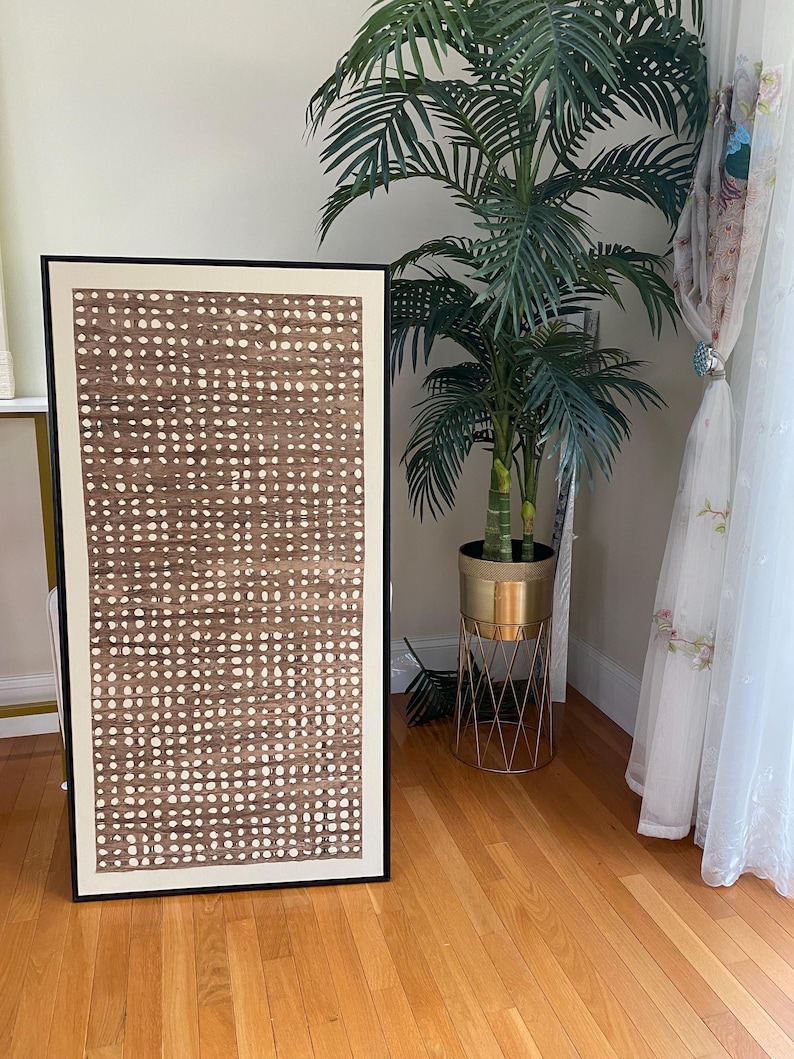 Pode incluir: Uma obra de arte abstrata emoldurada com um padr&atilde;o de pontos marrons e brancos. A obra de arte est&aacute; em uma moldura preta e encostada em uma parede. Uma palmeira alta em vaso e um suporte de planta dourado est&atilde;o ao fundo.