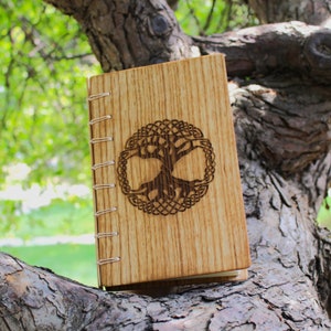 May include: A wooden journal with a Tree of Life design carved into the cover. The journal is light brown with a vertical wood grain pattern. The pages are bound with cream-colored thread.