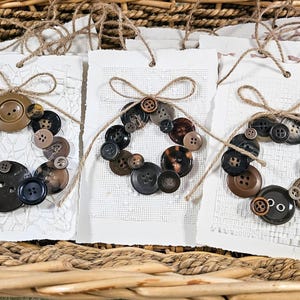 May include: Three handmade gift tags with button wreath designs. Each tag features a wreath made of various brown and black buttons, tied with twine bows. The tags are displayed in a woven basket.