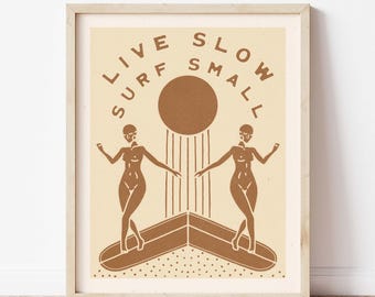 Può includere: Una stampa artistica incorniciata con sfondo beige e illustrazioni marroni. La stampa presenta due figure che fanno surf su una tavola da surf, con le parole "LIVE SURF SLOW SMALL" in un semicerchio sopra un sole marrone.