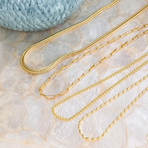 May include: A collection of gold necklaces with various chain styles, including a herringbone, paperclip, and beaded design. The necklaces are displayed on a marble surface, with a blue woven basket in the background.