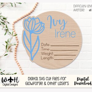 Könnte beinhalten: Eine hellblaue Blume mit Stiel und Blättern befindet sich auf einem runden Holzschild mit dem Namen "Ivy Irene" und Platz zum Ausfüllen von Datum, Uhrzeit, Gewicht und Länge. Das Schild befindet sich auf einem weißen Holzgrund.