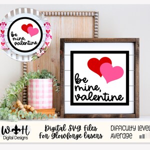 Könnte beinhalten: Eine digitale SVG-Datei für ein Valentinstagsschild mit dem Text "be mine, valentine" und zwei überlappenden Herzen, einem roten und einem rosa. Das Schild ist in einem dunklen Holzrahmen gerahmt und steht auf einem Regal mit einer Pflanze und einem Teller.