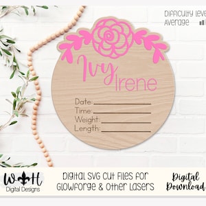 Könnte beinhalten: Ein rosa-weißes Baby-Ankündigungs-Schild aus Holz mit einem floralen Design und dem Namen "Ivy Irene" in rosa Schrift. Das Schild hat Platz zum Eintragen von Datum, Uhrzeit, Gewicht und Größe des Babys.
