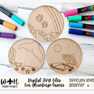May include: Three unfinished wooden coasters with different designs: a rainbow with butterflies, a beach scene with an umbrella, and a seashell and crab design. The coasters are ready to be painted with markers.
