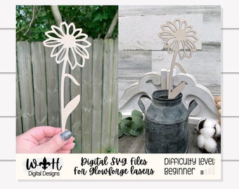 May include: A wooden cutout of a daisy flower with a stem and leaves. The flower is in a rustic setting with a metal pitcher and cotton balls. The text "Digital SVG files for Glowforge lasers" and "Difficulty level: beginner" is visible in the image.