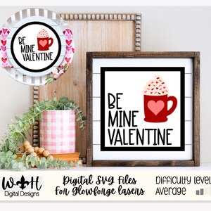 Könnte beinhalten: Eine digitale SVG-Datei für ein Valentinstagsschild mit der Aufschrift "Be Mine Valentine" und einer roten Tasse mit einem Herzen und Schlagsahne oben drauf. Das Schild ist in einem braunen Rahmen und steht auf einer weißen Holzoberfläche.