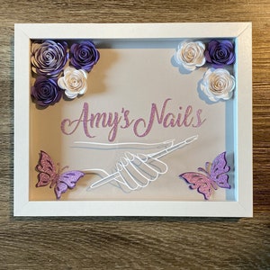 May include: A white frame with a light brown background. The frame has a hand drawn image of a hand holding a nail file. The text "Amy's Nails" is written in pink glitter letters. The frame is decorated with purple and white paper flowers and two pink glitter butterflies.