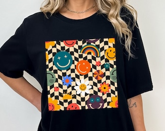 Puede incluir: Camiseta negra de gran tamaño con un estampado gráfico de caras sonrientes de colores, flores y otros diseños retro sobre un fondo a cuadros.