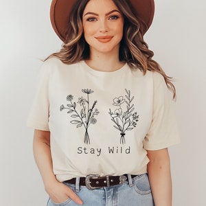 A short sleeve t-shirt with a minimalist floral graphic of two wildflower bouquets. Under it says "Stay Wild". Entire design is in black. Shirt is the Bella Canvas 3001 unisex jersey tee. Very high quality and soft.