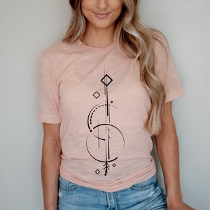 Pode incluir: Uma mulher vestindo uma camiseta rosa claro com um design geom&eacute;trico preto. O design apresenta uma forma de diamante, c&iacute;rculos e linhas.
