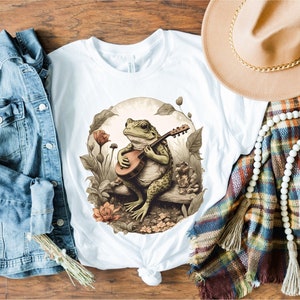 May include: A white t-shirt with a graphic of a frog playing a guitar in a forest setting. The frog is green and brown with a white belly. The guitar is brown and has a white pickguard. The forest is green and brown with flowers and leaves.