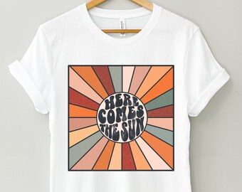 May include: White t-shirt with a retro-style graphic design featuring a sunburst pattern in shades of orange, brown, and green. The graphic says "Here Comes The Sun" in a groovy font.