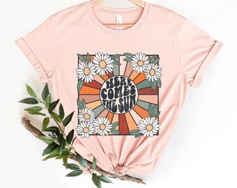 May include: A pink t-shirt with a retro-style graphic featuring a sun with the text "Here Comes The Sun" surrounded by daisies.