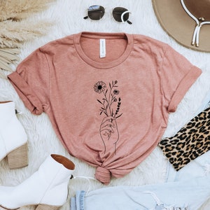 May include: A dusty rose t-shirt with a black line drawing of a hand holding a bouquet of flowers.