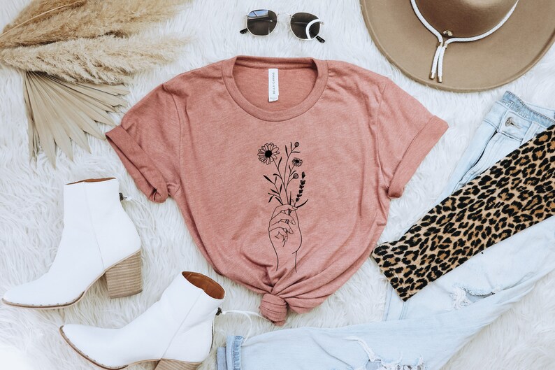 May include: A dusty rose t-shirt with a black line drawing of a hand holding a bouquet of flowers.