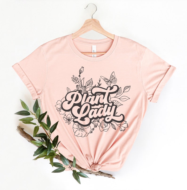 A short sleeve t shirt that says "Plant Lady" with leaves and flowers behind it. The shirt is Bella and Canvas 3001 unisex jersey tee. It is very soft and high quality and comes in different colors.