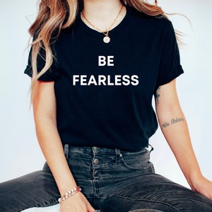 May include: A black t-shirt with the text "BE FEARLESS" printed in white on the front. The shirt is being worn by a person with long blonde hair and a gold necklace.