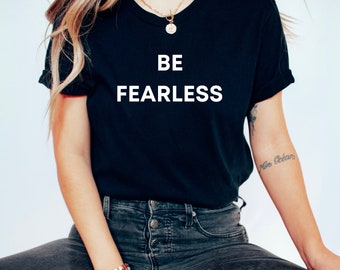 Puede incluir: Una camiseta negra con el texto "BE FEARLESS" impreso en blanco en la parte delantera. La camiseta la lleva puesta una persona con el pelo largo y rubio y un collar de oro.