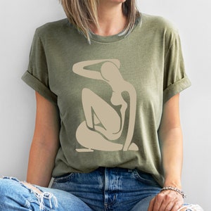 May include: A green t-shirt with a beige abstract figure design. The figure is stylized and resembles a person sitting with their arm raised above their head.