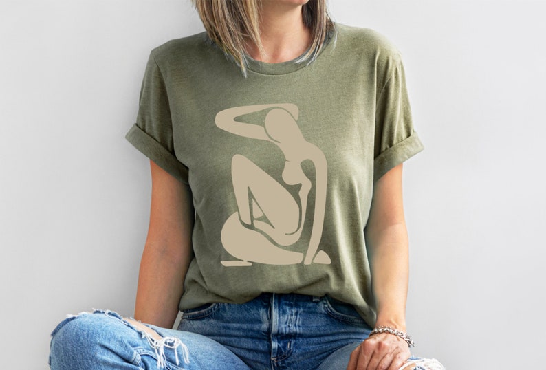 May include: A green t-shirt with a beige abstract figure design. The figure is stylized and resembles a person sitting with their arm raised above their head.