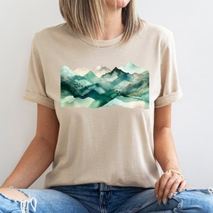 Puede incluir: Una camiseta beige con un dise&ntilde;o de acuarela de monta&ntilde;as verdes en la parte delantera.