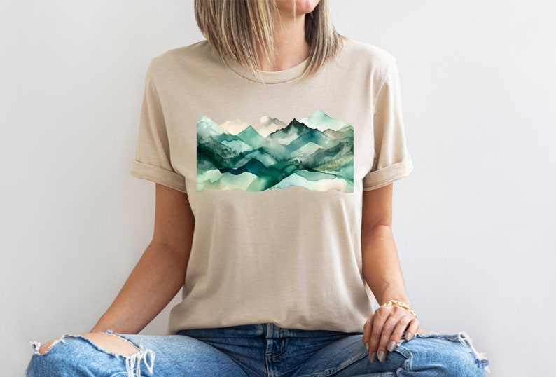 Puede incluir: Una camiseta beige con un dise&ntilde;o de acuarela de monta&ntilde;as verdes en la parte delantera.