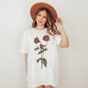 May include: A white t-shirt with a floral print of burgundy flowers and green leaves. The shirt has a relaxed fit and a crew neck.