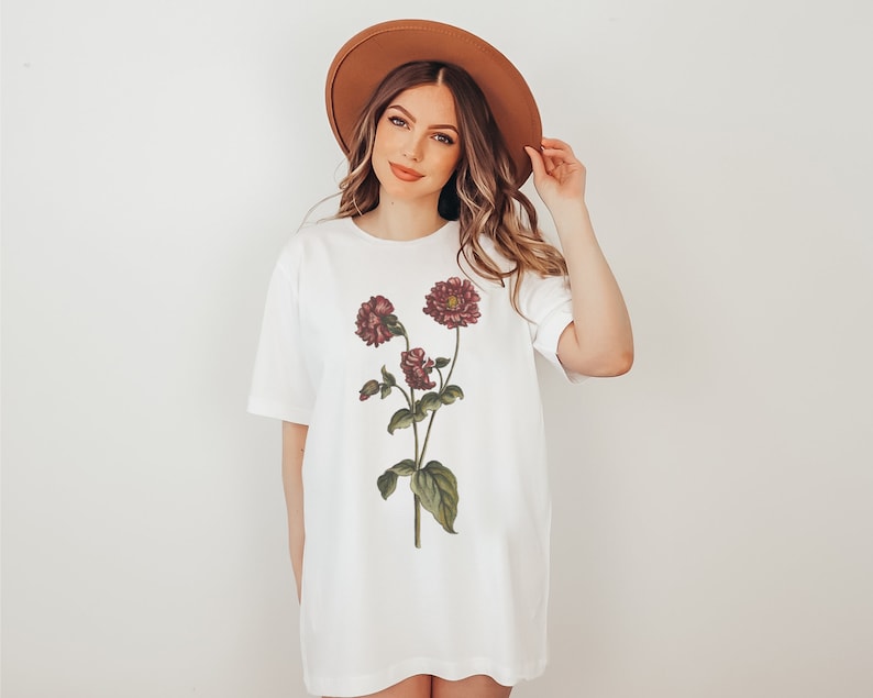 May include: A white t-shirt with a floral print of burgundy flowers and green leaves. The shirt has a relaxed fit and a crew neck.