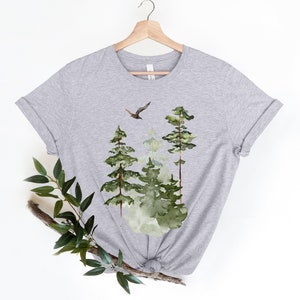 May include: Gray t-shirt with a watercolor design of a forest scene with three trees and a bird flying overhead.