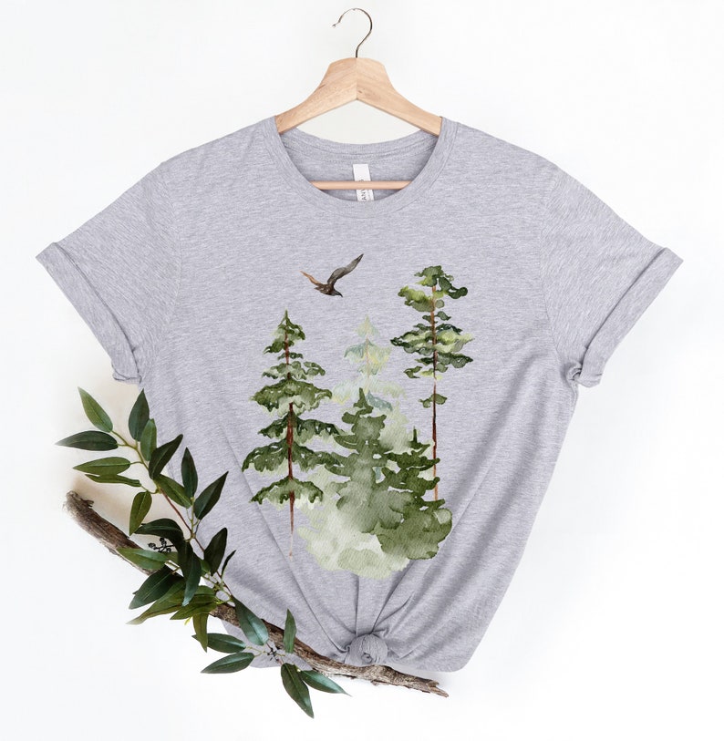 May include: Gray t-shirt with a watercolor design of a forest scene with three trees and a bird flying overhead.