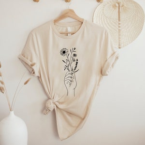 May include: A beige t-shirt with a black line drawing of a hand holding a bouquet of flowers.