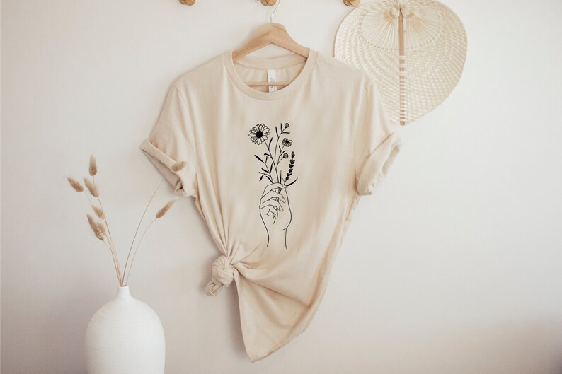 May include: A beige t-shirt with a black line drawing of a hand holding a bouquet of flowers.