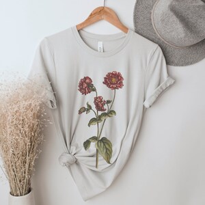 May include: A light gray t-shirt with a floral print of red flowers on a green stem. The shirt is folded and displayed on a hanger.