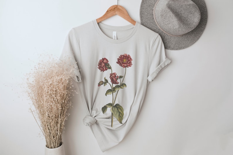 May include: A light gray t-shirt with a floral print of red flowers on a green stem. The shirt is folded and displayed on a hanger.