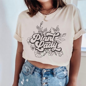May include: A cream-colored t-shirt with a black graphic design of flowers and the text "Plant Lady" in a retro font.