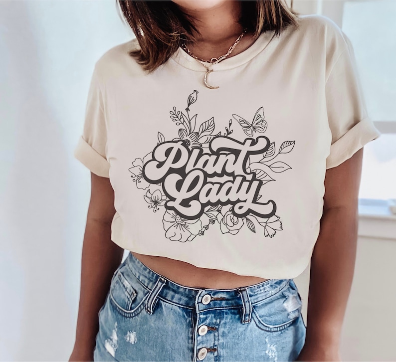 May include: A cream-colored t-shirt with a black graphic design of flowers and the text "Plant Lady" in a retro font.