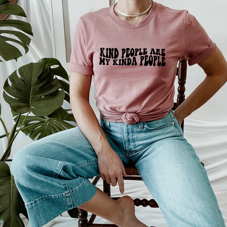 May include: A woman wearing a dusty rose t-shirt with the text "Kind People Are My Kinda People" in black retro font. She is wearing blue jeans and sitting on a wooden chair.