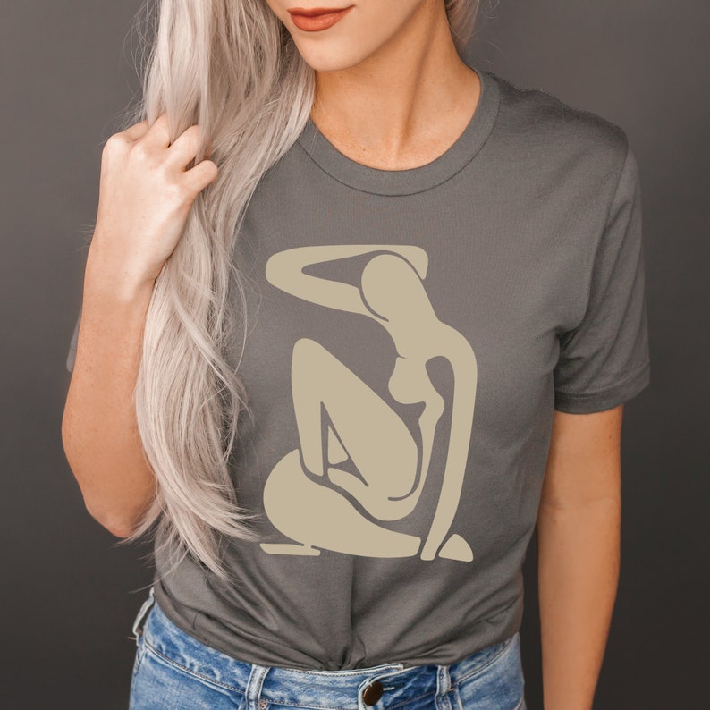 May include: A charcoal grey t-shirt with a beige abstract figure design. The figure is stylized and has a rounded, organic shape.