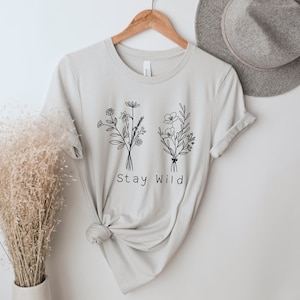 May include: A light gray t-shirt with a black line drawing of two bouquets of flowers and the text "Stay Wild".