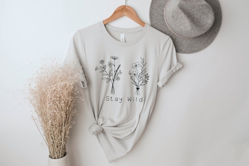 May include: A light gray t-shirt with a black line drawing of two bouquets of flowers and the text "Stay Wild".